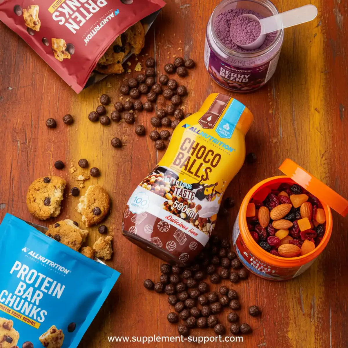 All Nutrition Choco Balls Sauce neben Proteinriegeln, Cookies und Beerenmischung auf Holzuntergrund.
