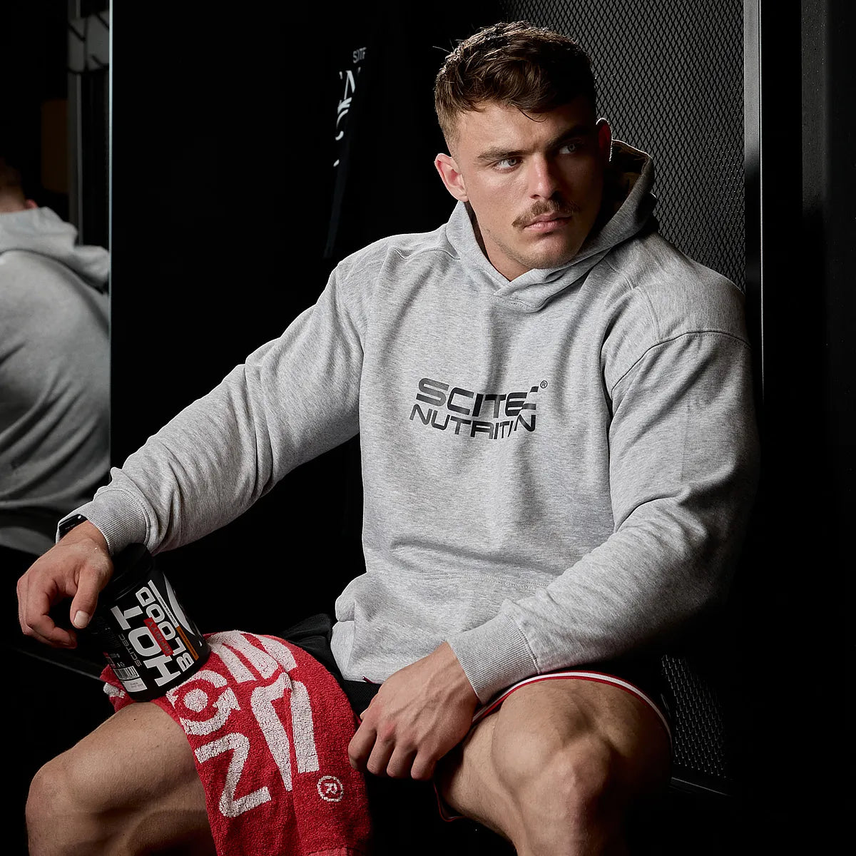 Sportler mit grauem Hoodie, hält Scitec Nutrition Hot Blood Booster im Fitnessstudio